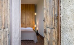 Cozy bedroom interior with a wooden wall and a soft bedspread, viewed through rustic wooden doors. The room features a bedside lamp and warm lighting, creating a welcoming atmosphere. Ideal for showcasing comfortable hotel accommodations or home decor inspiration.