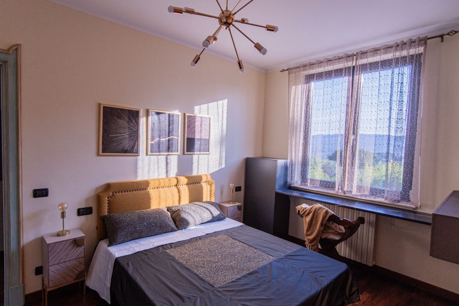 Modern bedroom interior featuring a stylish bed with decorative pillows, framed artwork on the wall, a cozy chair, and a window with sheer curtains allowing natural light to illuminate the space. | Only on SopranoVillas