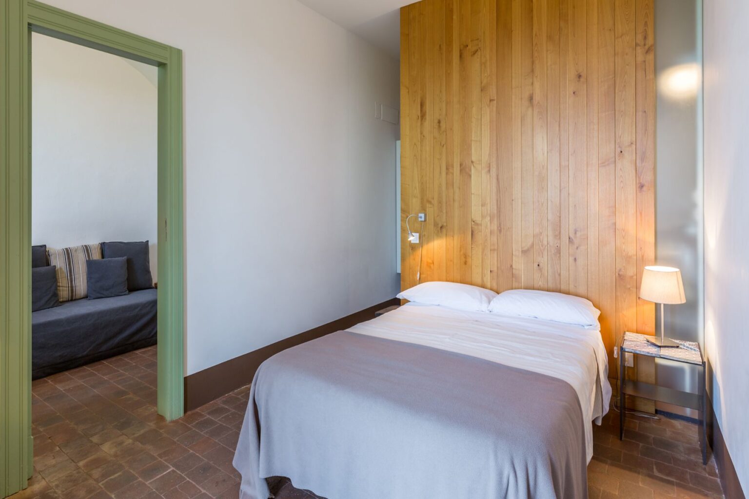 Cozy bedroom featuring a wooden accent wall, a comfortable double bed with white linens, and a stylish bedside lamp. A doorway leads to a separate sitting area with a sofa, enhancing the room's spacious feel. The warm color palette and natural light create a welcoming atmosphere. | Only on SopranoVillas