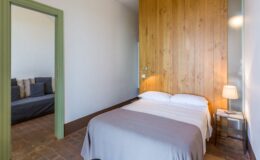 Cozy bedroom featuring a wooden accent wall, a comfortable double bed with white linens, and a stylish bedside lamp. A doorway leads to a separate sitting area with a sofa, enhancing the room's spacious feel. The warm color palette and natural light create a welcoming atmosphere.