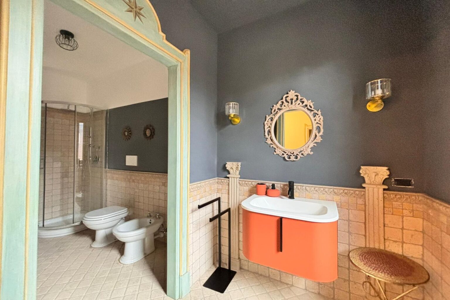 Modern bathroom interior featuring a stylish orange sink, decorative mirror, and elegant wall sconces. The space includes a shower area, bidet, and unique tile work, creating a blend of contemporary and classic design elements. | Only on SopranoVillas