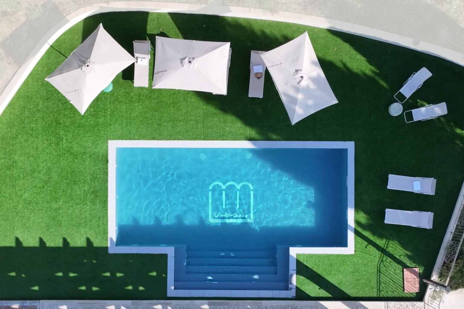 Aerial view of a modern swimming pool surrounded by lush green artificial grass, featuring sun loungers and large umbrellas for shade, perfect for relaxation and outdoor gatherings. The pool displays a logo at the bottom, adding a stylish touch to the serene setting. | Only on SopranoVillas