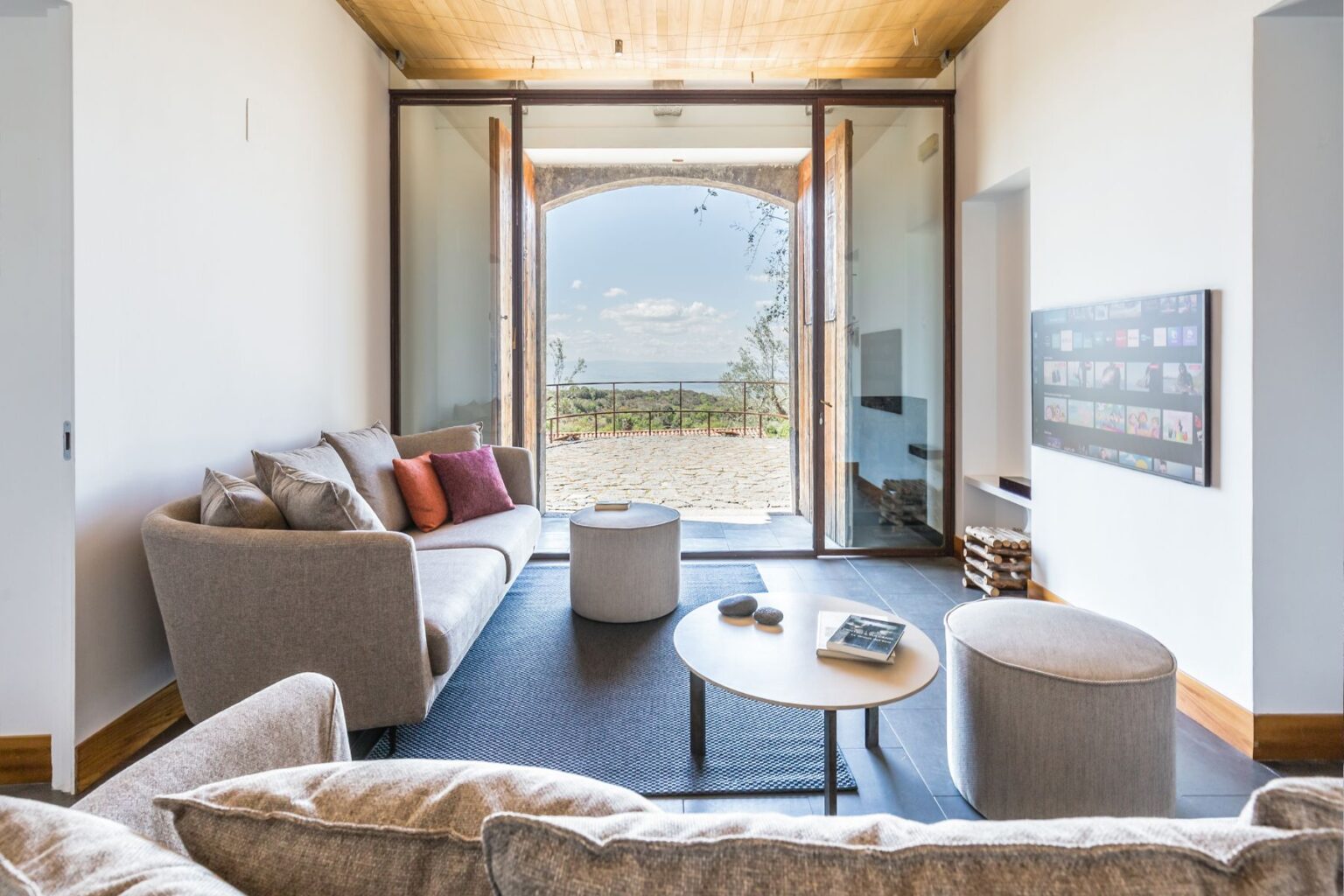 Modern living room with a cozy gray sofa and colorful cushions, featuring a round coffee table and a flat-screen TV. Large glass doors open to a scenic outdoor view, blending indoor and outdoor spaces. Natural light illuminates the room, enhancing the contemporary design. | Only on SopranoVillas