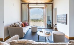 Modern living room with a cozy gray sofa and colorful cushions, featuring a round coffee table and a flat-screen TV. Large glass doors open to a scenic outdoor view, blending indoor and outdoor spaces. Natural light illuminates the room, enhancing the contemporary design.