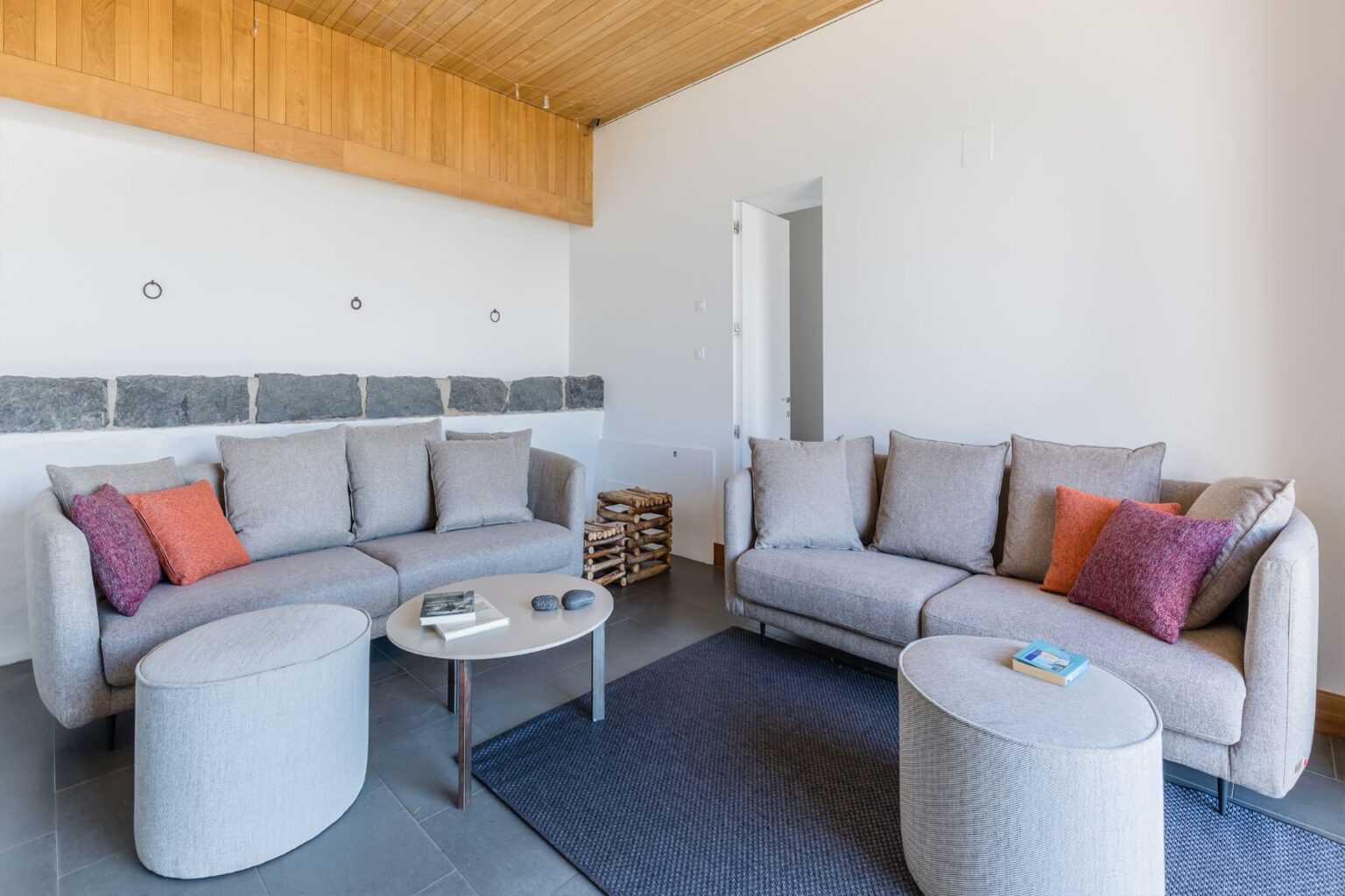 Modern living room with gray sofas, decorative pillows, a round coffee table, and a textured rug. Features wooden ceiling and stone wall accents, creating a cozy and stylish atmosphere. | Only on SopranoVillas