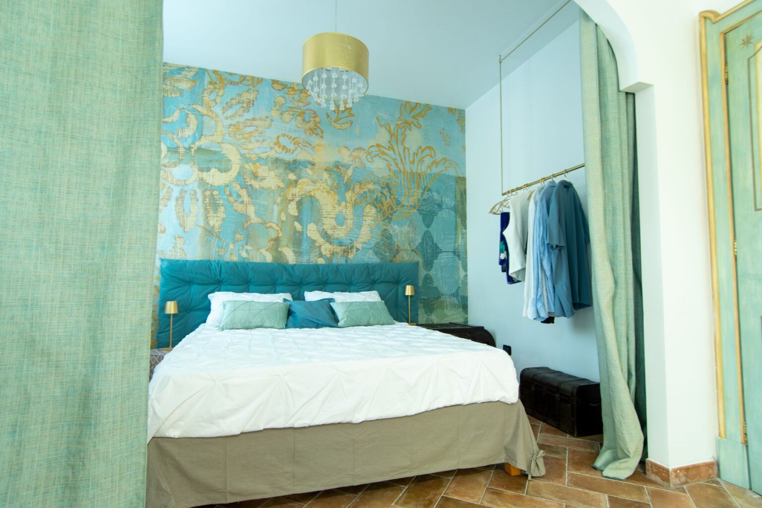 Modern bedroom featuring a teal upholstered headboard, a stylish light fixture, and a decorative wall with gold accents. The room includes a neatly made bed with white and teal bedding, a cozy atmosphere, and a clothing rack displaying shirts. The floor is adorned with warm-toned tiles, enhancing the inviting design. | Only on SopranoVillas