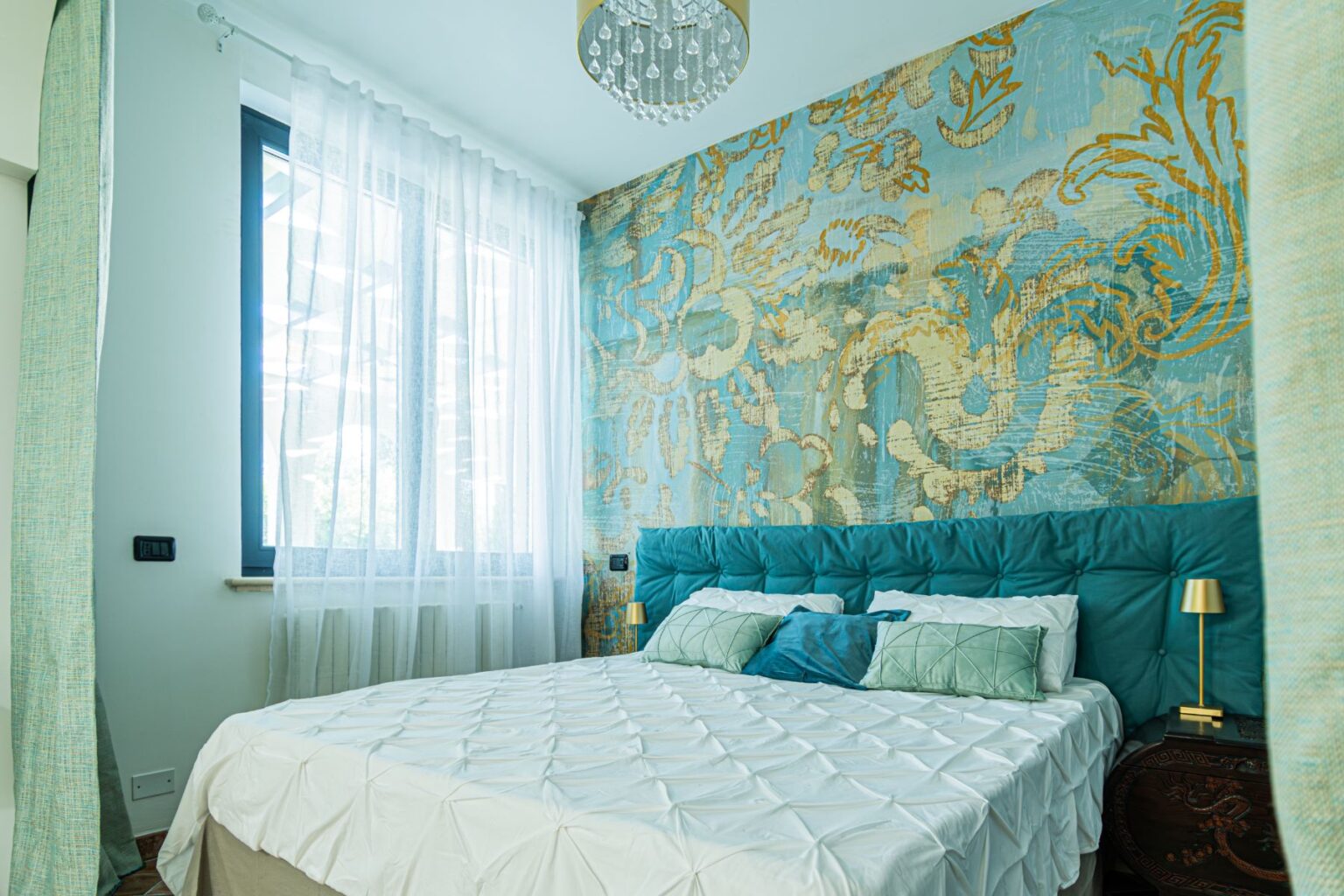 Cozy bedroom featuring a king-sized bed with a textured white duvet, teal upholstered headboard, and decorative pillows. The room is adorned with a vibrant blue and gold patterned accent wall, sheer white curtains allowing natural light, and a modern chandelier. A stylish bedside table with a gold lamp completes the inviting ambiance. | Only on SopranoVillas
