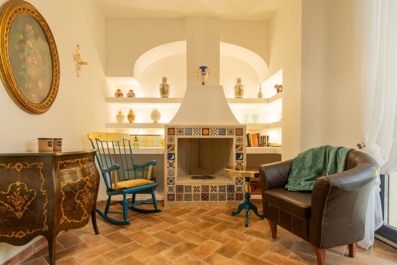 Cozy living room featuring a traditional fireplace adorned with colorful tiles, a blue rocking chair, and a brown leather armchair. Decorative shelves display various ceramic vases, while the warm tiled floor and soft lighting create an inviting atmosphere. | Only on SopranoVillas