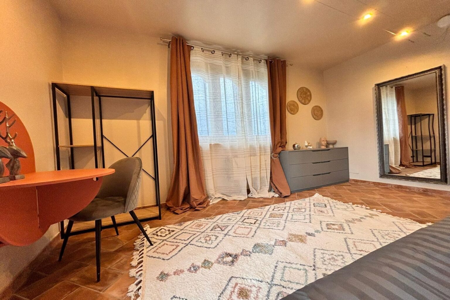 Cozy bedroom interior featuring a decorative rug, a stylish gray dresser, a modern desk with a chair, and large windows dressed with sheer and brown curtains, complemented by wall decor and a full-length mirror. | Only on SopranoVillas