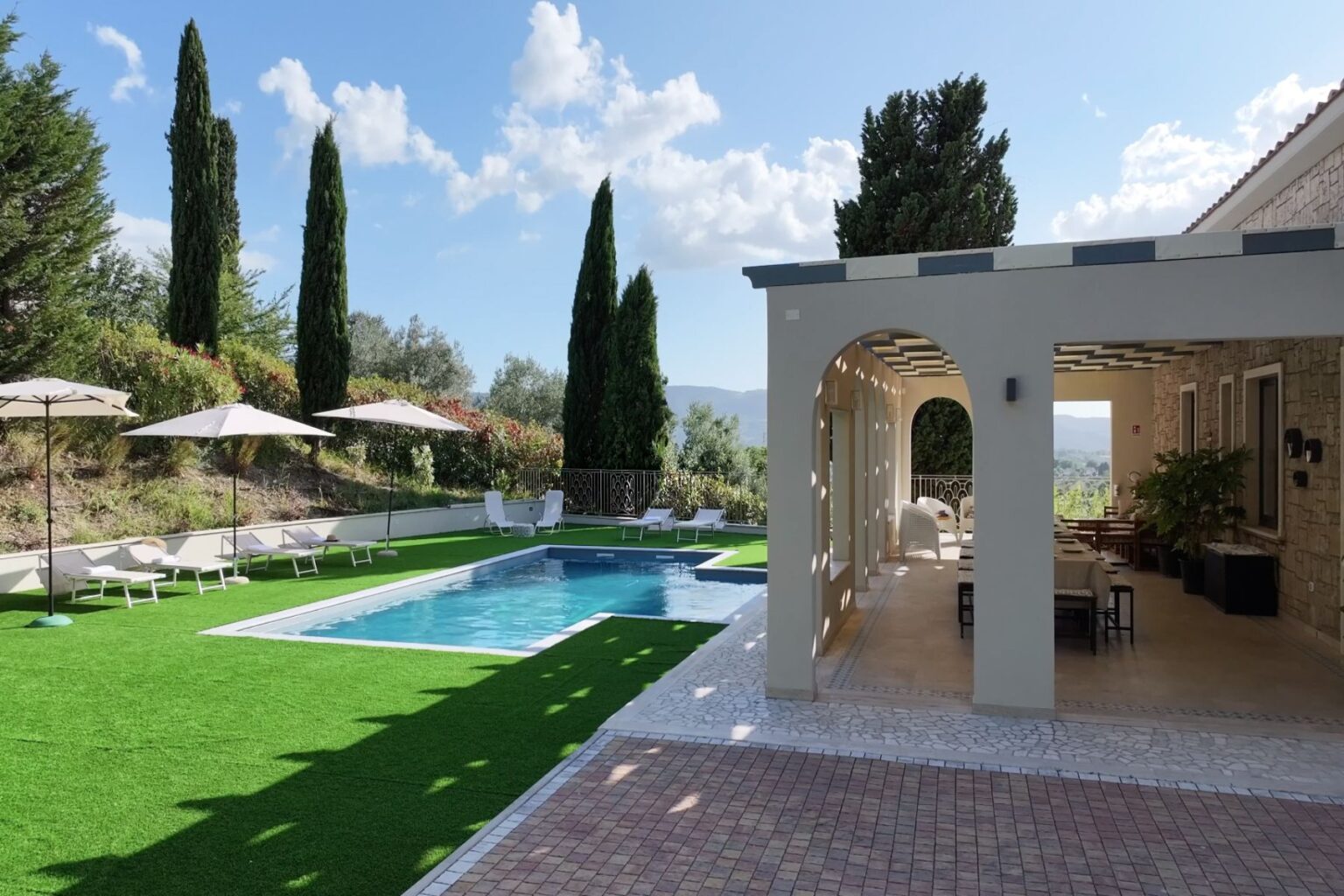 Luxurious villa with a swimming pool, surrounded by lush greenery and tall cypress trees. Sun loungers and umbrellas line the pool area, providing a relaxing outdoor space. The villa features a spacious terrace and elegant architecture, perfect for leisure and entertaining. | Only on SopranoVillas
