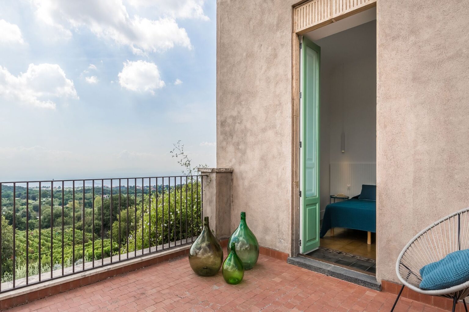 Contemporary balcony view featuring green decorative glass bottles, a light-colored wall, and an open door leading to a stylish bedroom with a blue bedspread. Lush greenery and a scenic landscape are visible in the background under a partly cloudy sky. | Only on SopranoVillas