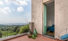 Contemporary balcony view featuring green decorative glass bottles, a light-colored wall, and an open door leading to a stylish bedroom with a blue bedspread. Lush greenery and a scenic landscape are visible in the background under a partly cloudy sky.