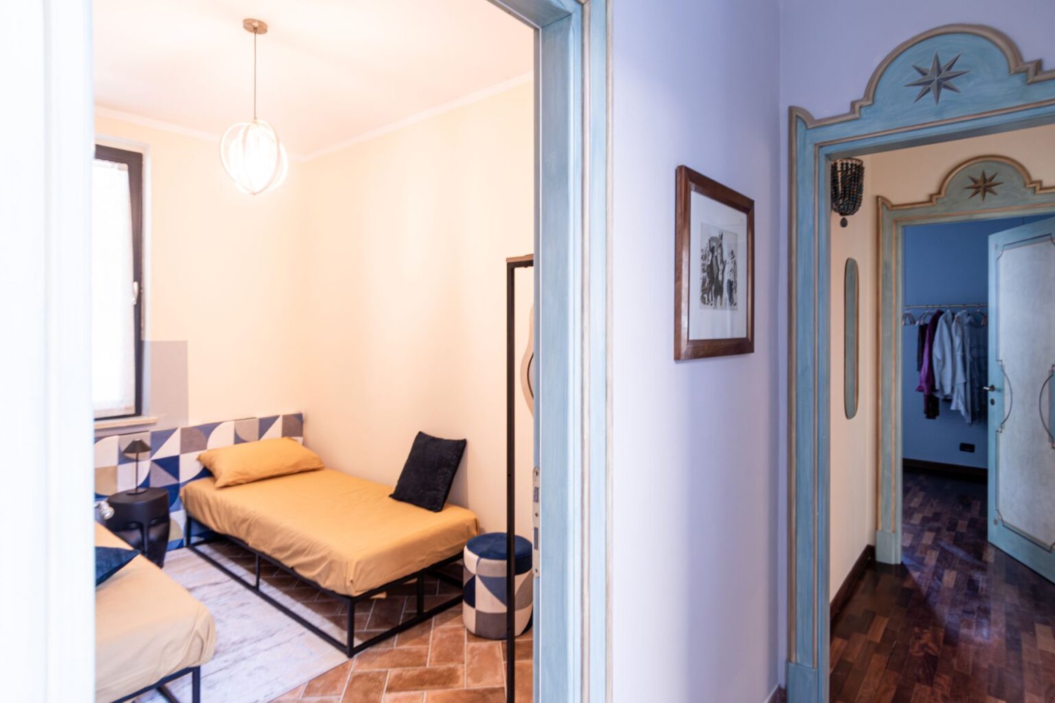 Cozy bedroom interior featuring two single beds with yellow bedding, decorative pillows, and modern lighting. The room has a stylish geometric tile accent wall and is connected to a hallway with wooden flooring and a glimpse of a closet. | Only on SopranoVillas