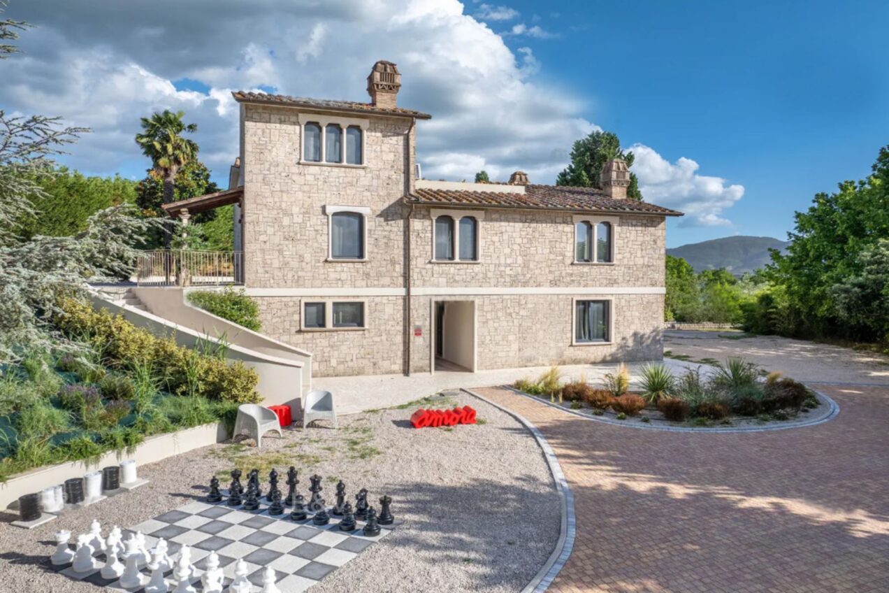 Stunning stone house with modern architecture, featuring a landscaped garden, outdoor chess set, and scenic mountain views under a bright blue sky. Perfect for luxury living or vacation rentals. | Only on SopranoVillas