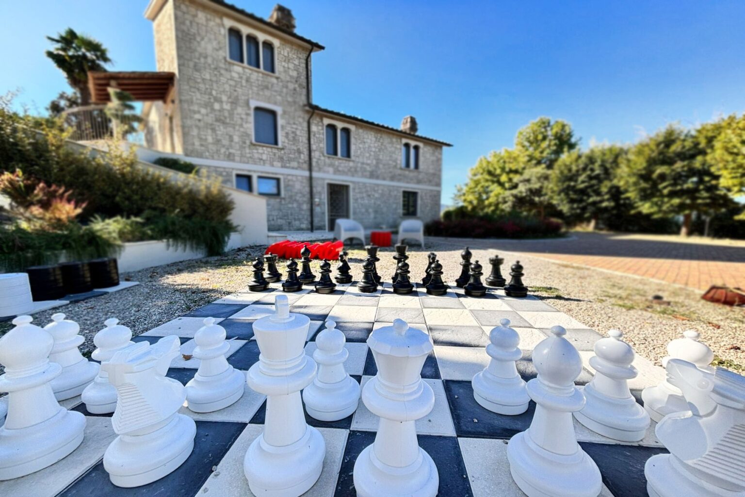 Outdoor giant chess set in front of a stone house, with white and black pieces arranged on a checkered board, surrounded by greenery and blue skies. | Only on SopranoVillas