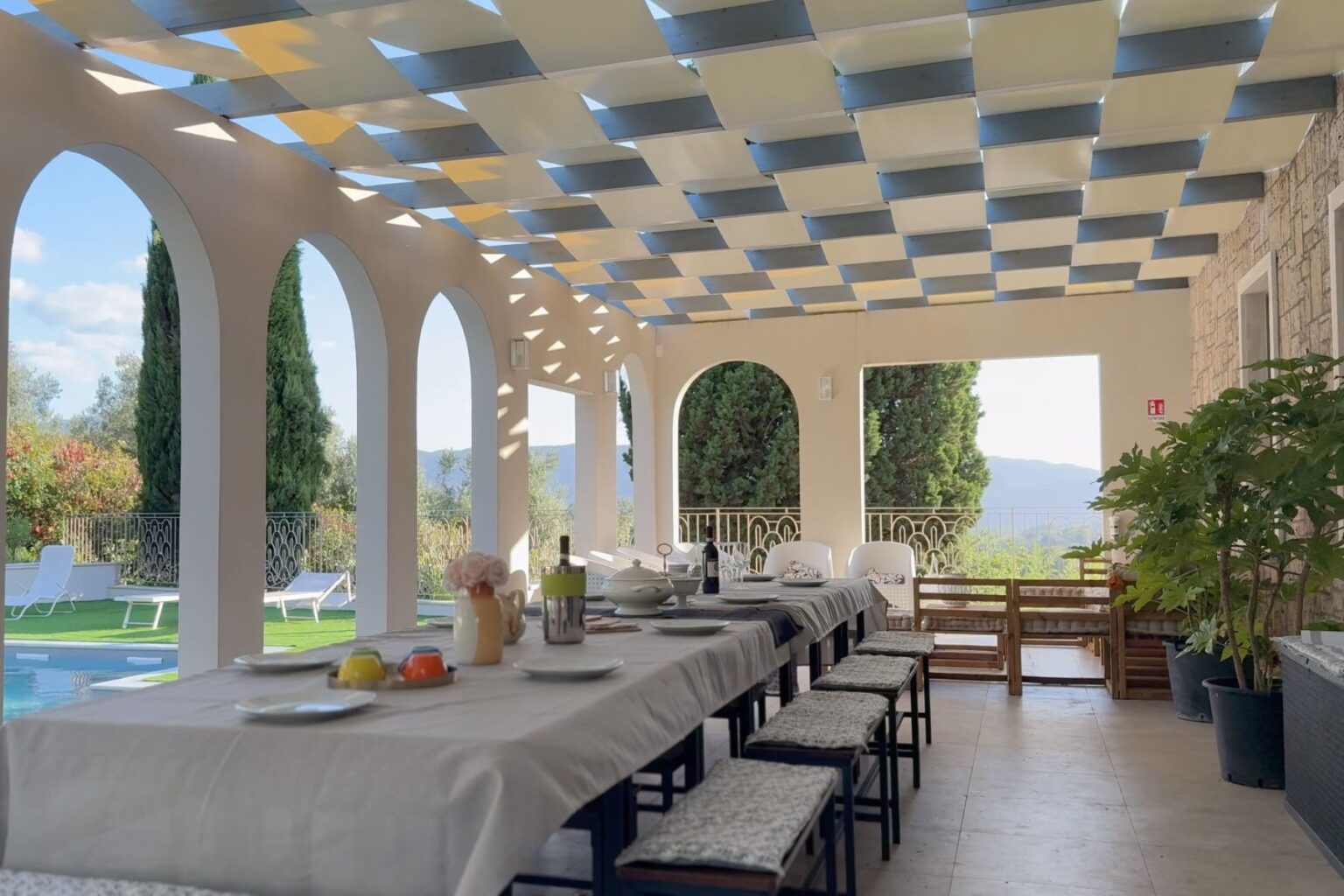Spacious outdoor dining area with a long table set for a meal, featuring a patterned pergola overhead, elegant arches, and views of a garden and swimming pool. Ideal for gatherings and al fresco dining. | Only on SopranoVillas