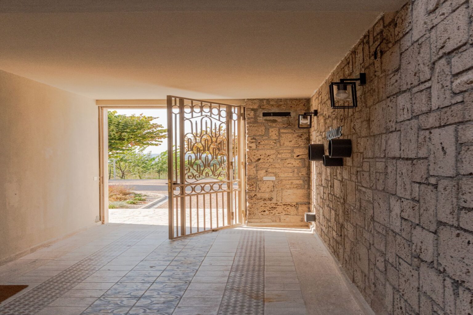 Open doorway leading to a bright outdoor space, featuring a decorative metal gate and a stone wall interior with wall-mounted lanterns. The tiled floor showcases intricate patterns, and green trees are visible outside, enhancing the inviting atmosphere. | Only on SopranoVillas