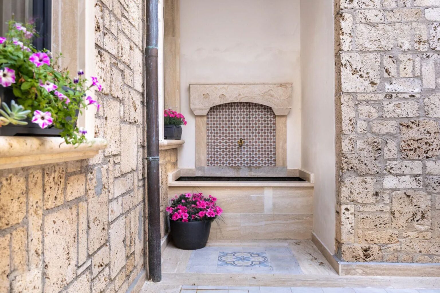 Charming courtyard featuring a decorative stone fountain with intricate tile work, surrounded by vibrant potted flowers and textured stone walls, creating a serene outdoor space. | Only on SopranoVillas