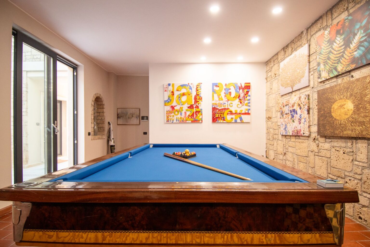 Interior view of a stylish game room featuring a blue pool table, decorative wall art, and large windows allowing natural light. The room combines modern and artistic elements, creating an inviting atmosphere for leisure activities. | Only on SopranoVillas
