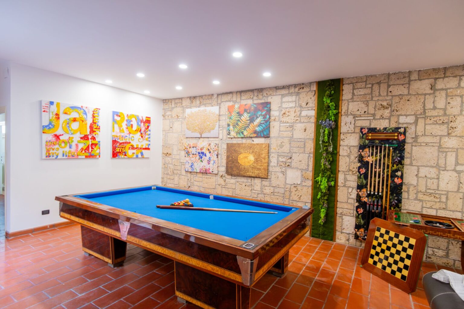 Interior view of a stylish game room featuring a blue pool table, vibrant art pieces on the walls, and decorative stone accents. The space includes a checkered game board and greenery, creating a modern yet inviting atmosphere for leisure activities. | Only on SopranoVillas