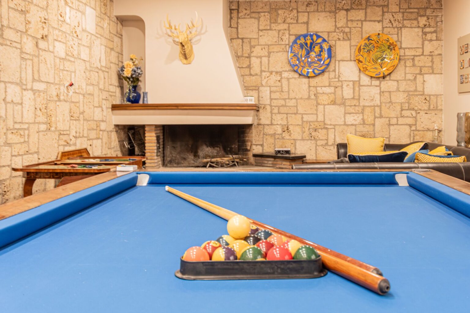 Interior view of a cozy game room featuring a blue pool table with a set of colorful billiard balls arranged in a triangle. The stone wall backdrop includes decorative plates and a mounted deer head, while a fireplace and comfortable seating area with yellow cushions complete the inviting atmosphere. | Only on SopranoVillas