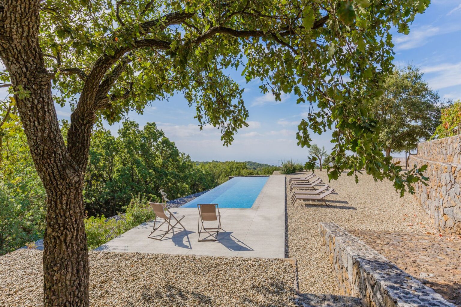 Serene outdoor pool area surrounded by lush greenery, featuring a modern infinity pool, sun loungers, and a shaded tree, perfect for relaxation and leisure. | Only on SopranoVillas