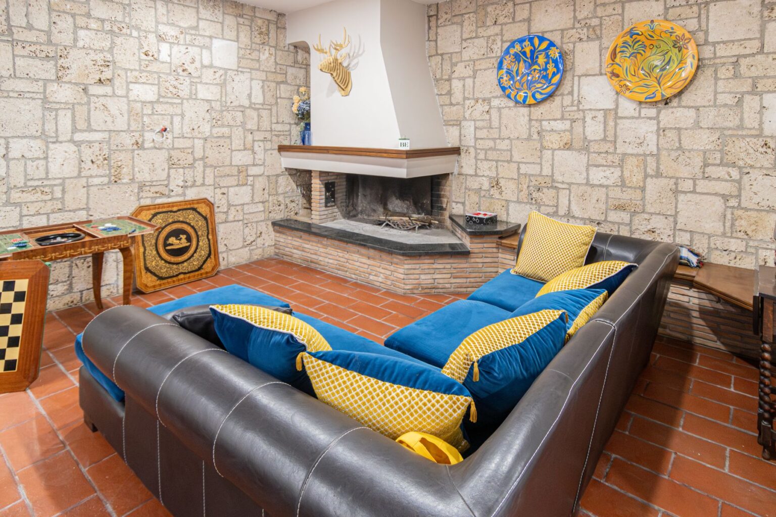 Cozy living room featuring a dark leather sectional sofa with vibrant blue and yellow cushions, a stone fireplace, and decorative plates on the wall. A wooden game table with a checkerboard design is also visible, enhancing the inviting atmosphere. | Only on SopranoVillas