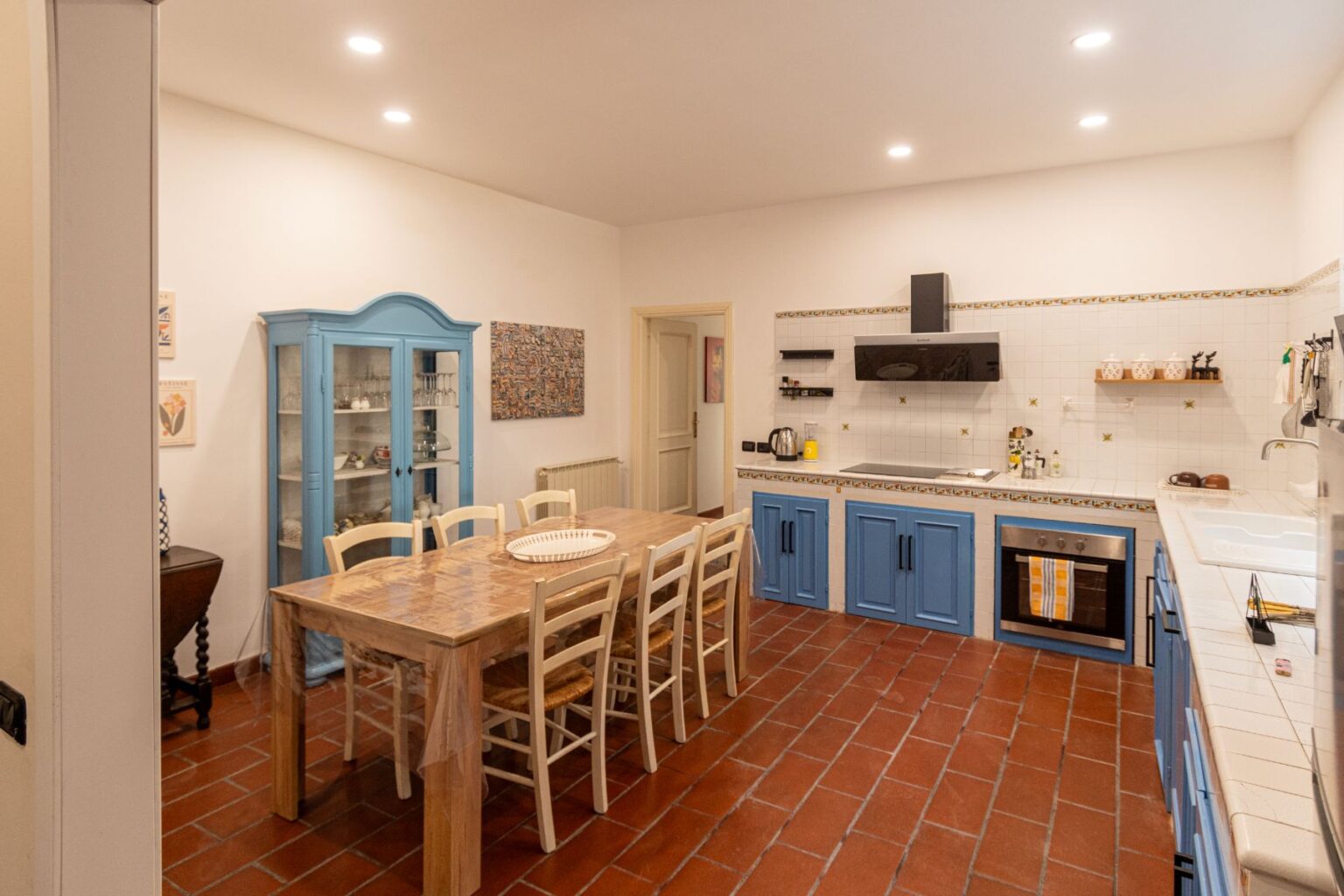 Spacious kitchen featuring blue cabinetry, a wooden dining table with eight chairs, and modern appliances. The room is illuminated with recessed lighting and showcases a decorative blue cabinet and wall art. The flooring consists of terracotta tiles, enhancing the warm ambiance. | Only on SopranoVillas