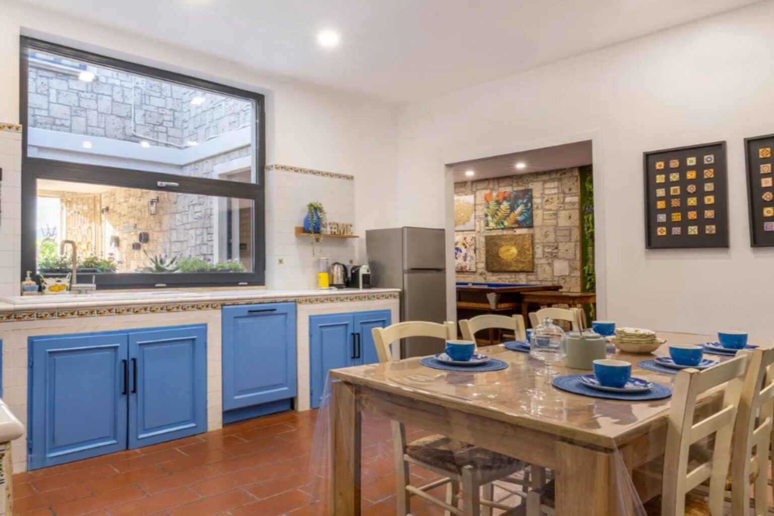 Bright and modern kitchen featuring blue cabinets, a wooden dining table set for six, and large windows that open to a lush courtyard. The space includes a refrigerator, coffee maker, and decorative artwork on the walls, creating a warm and inviting atmosphere. | Only on SopranoVillas