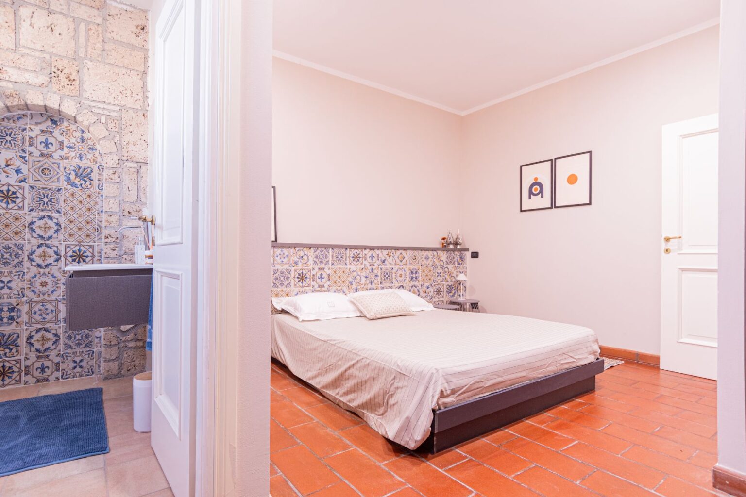 Cozy bedroom featuring a double bed with a patterned headboard, terracotta tile flooring, and a stylish wall with decorative tiles. The room includes framed artwork, a bedside table, and a glimpse of a modern bathroom area. Ideal for a relaxing stay. | Only on SopranoVillas