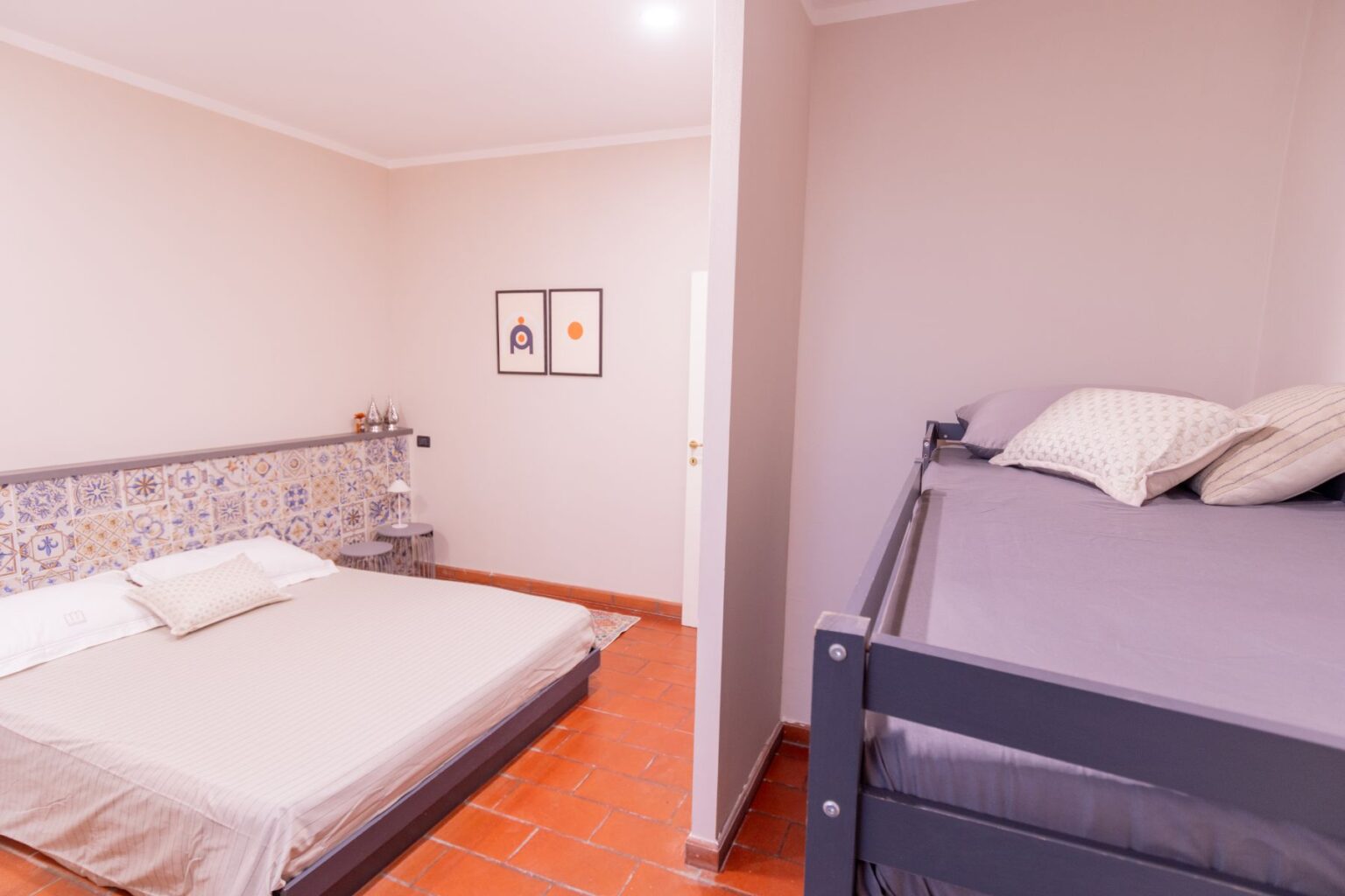 Cozy bedroom featuring a large double bed with a decorative tiled headboard and a bunk bed in a modern style. The room has soft lighting, warm terracotta flooring, and minimalist artwork on the walls, creating a welcoming atmosphere. Ideal for family stays or group accommodations. | Only on SopranoVillas