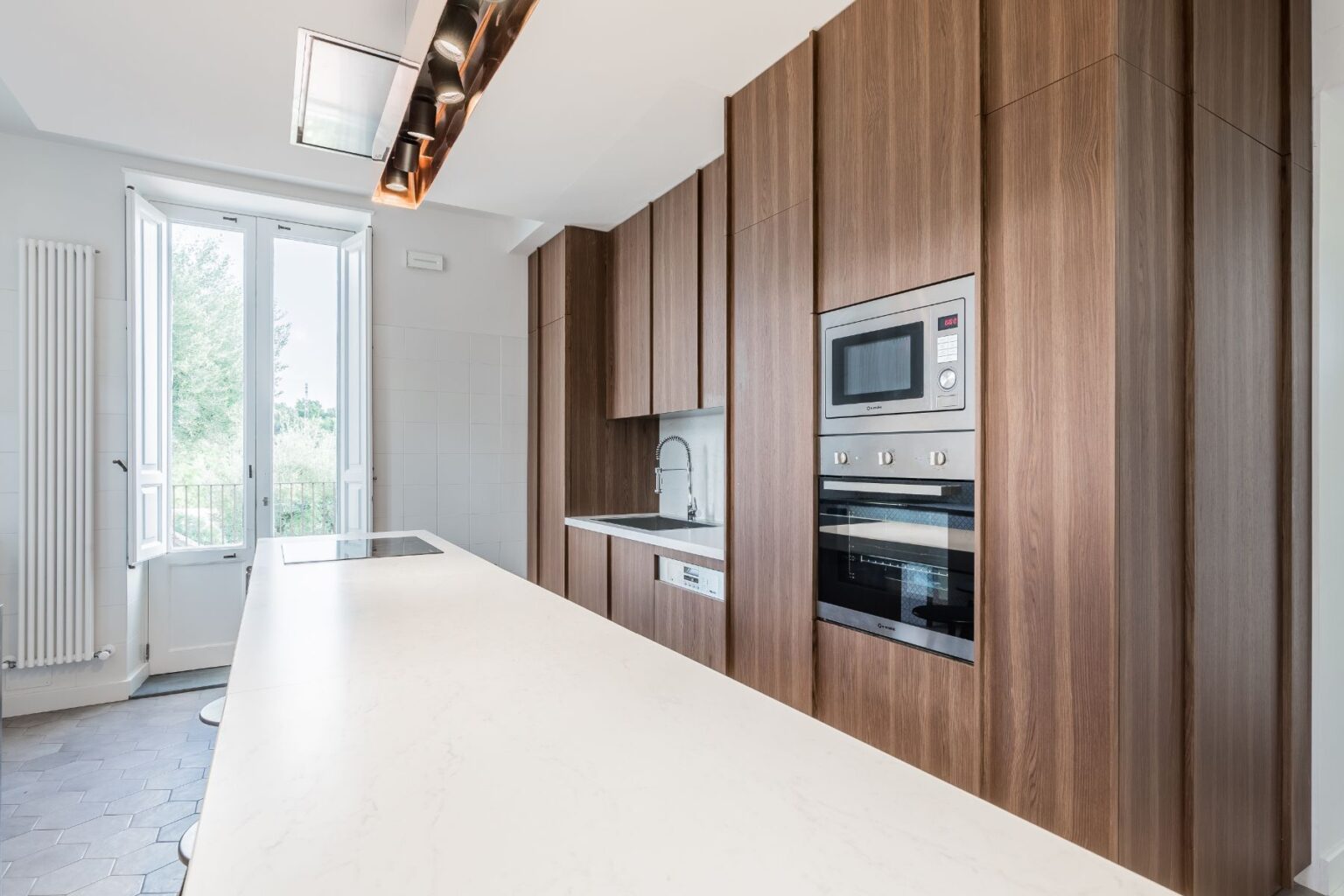 Modern kitchen with wooden cabinetry, sleek white countertops, and built-in appliances. Large windows provide natural light and a view of greenery outside, enhancing the spacious and contemporary design. | Only on SopranoVillas