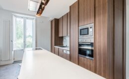 Modern kitchen with wooden cabinetry, sleek white countertops, and built-in appliances. Large windows provide natural light and a view of greenery outside, enhancing the spacious and contemporary design.