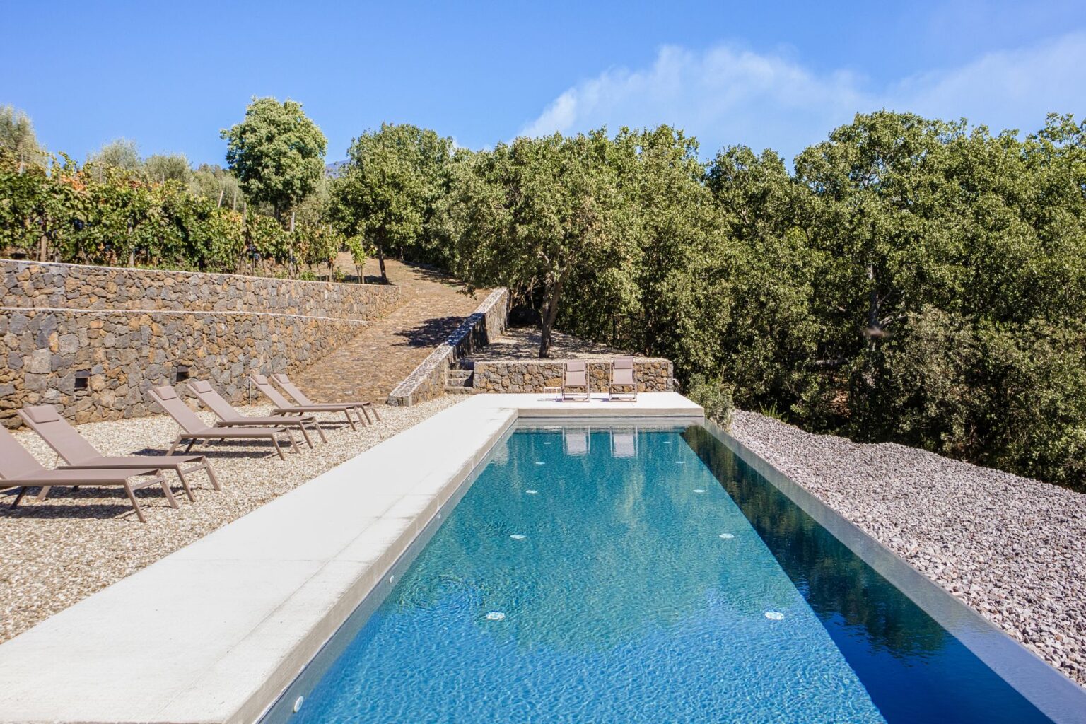 Modern infinity pool surrounded by lush greenery and stone pathways, featuring lounge chairs on a gravel patio, ideal for relaxation and outdoor enjoyment. | Only on SopranoVillas