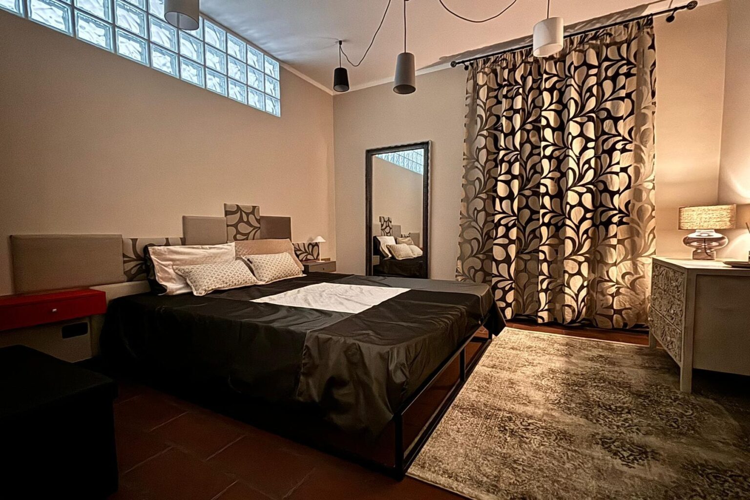 Modern bedroom interior featuring a black bed with decorative pillows, a large mirror, stylish curtains with a leaf pattern, and a bedside lamp. Natural light filters through a glass block window, enhancing the cozy atmosphere. A textured rug adds warmth to the space. | Only on SopranoVillas