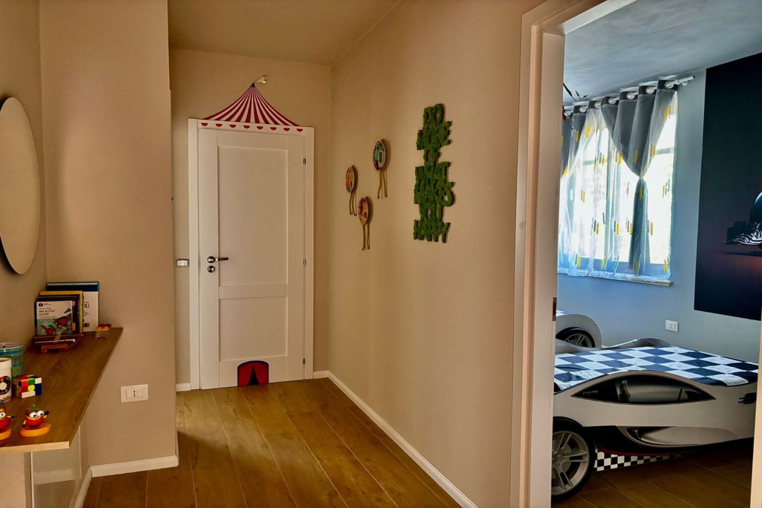 Bright and playful children's hallway featuring a white door with a circus-themed decoration, green wall art, and a wooden shelf displaying toys and books. The adjacent room is visible, showcasing a race car bed and cheerful window curtains. | Only on SopranoVillas