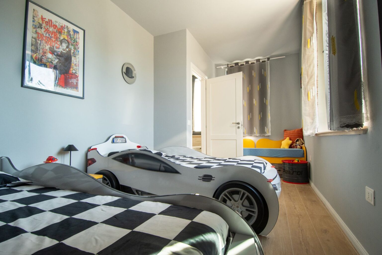Child's bedroom featuring two race car beds with black and white checkered bedding, a colorful seating area, and playful decor including framed art and curtains. The room is well-lit with natural light, creating a vibrant and fun atmosphere for kids. | Only on SopranoVillas