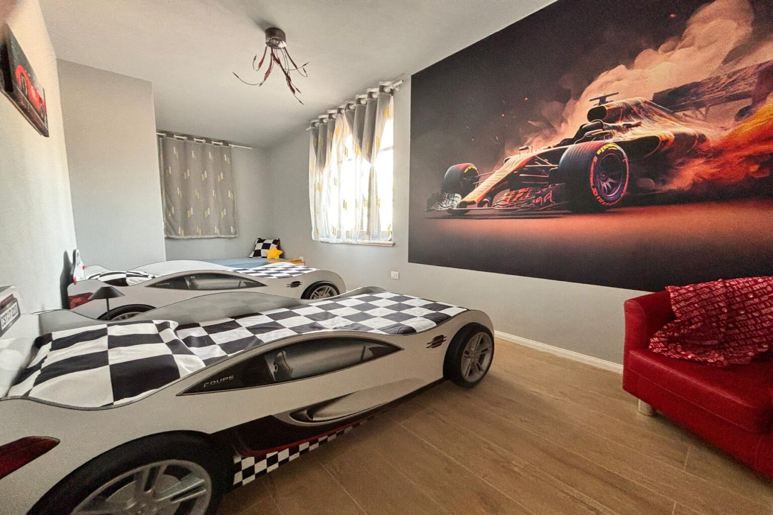 Children's bedroom featuring two race car beds with checkered bedding, a vibrant wall mural of a racing car, and bright curtains. The room is designed for young car enthusiasts, combining playful decor with a cozy atmosphere. | Only on SopranoVillas