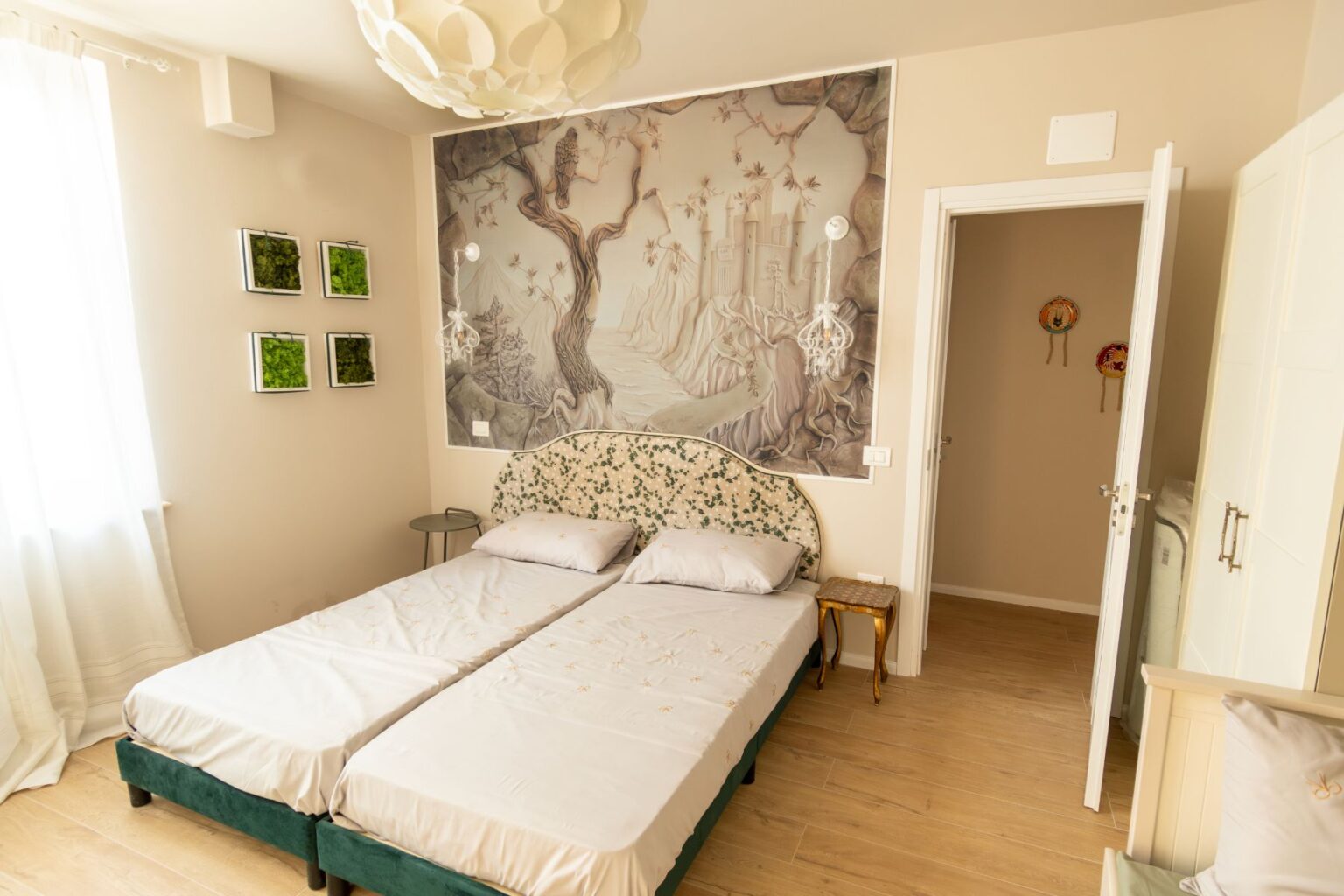 Cozy bedroom featuring twin beds with floral bedding, a large mural depicting a serene landscape on the wall, decorative green moss art frames, and a modern chandelier. Natural light streams in through sheer curtains, enhancing the warm, inviting atmosphere. | Only on SopranoVillas