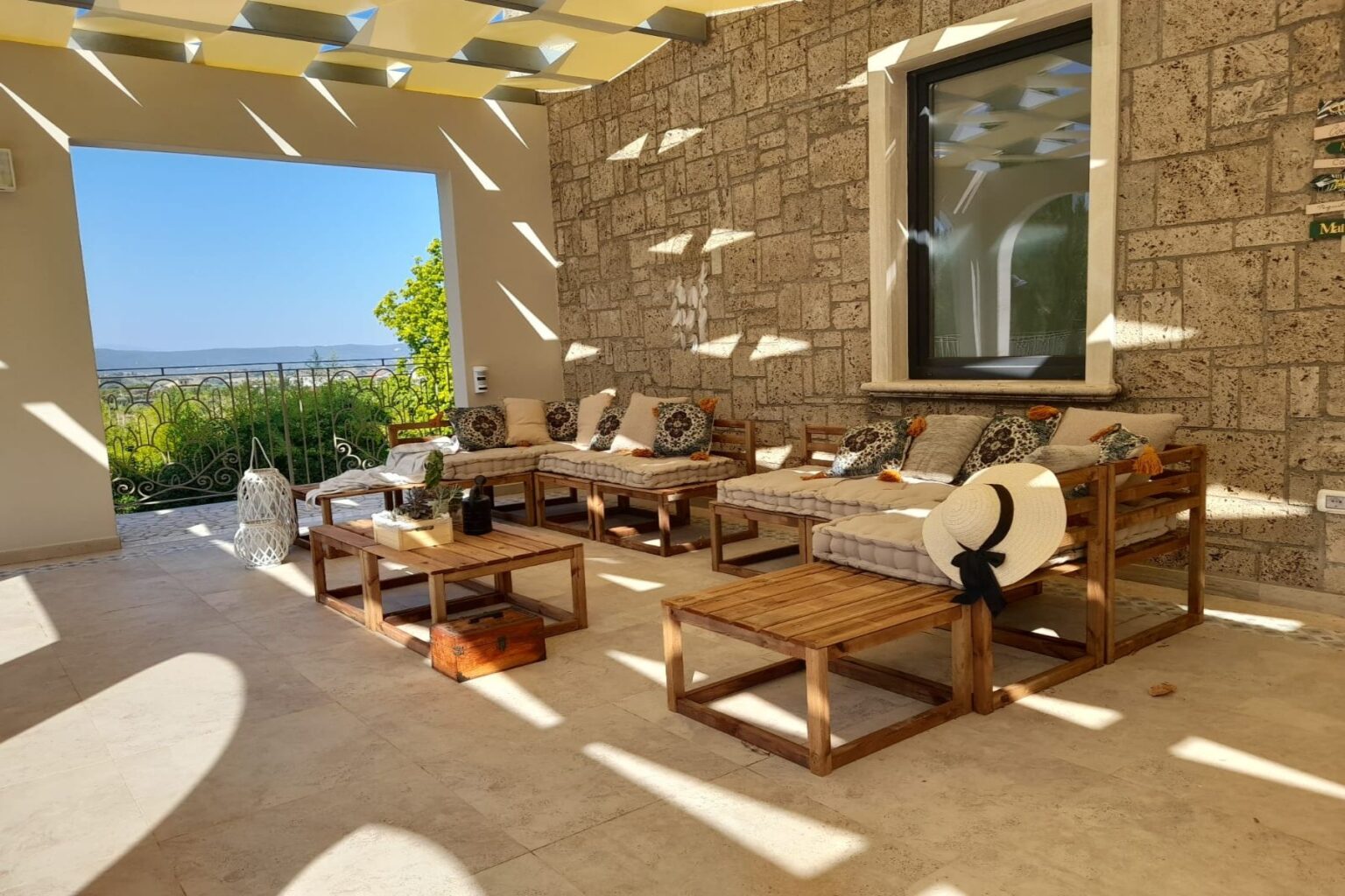 Cozy outdoor living space featuring wooden seating with cushions and decorative pillows, a view of greenery and hills through a large window, and natural light casting shadows on the stone wall. Ideal for relaxation and entertaining. | Only on SopranoVillas