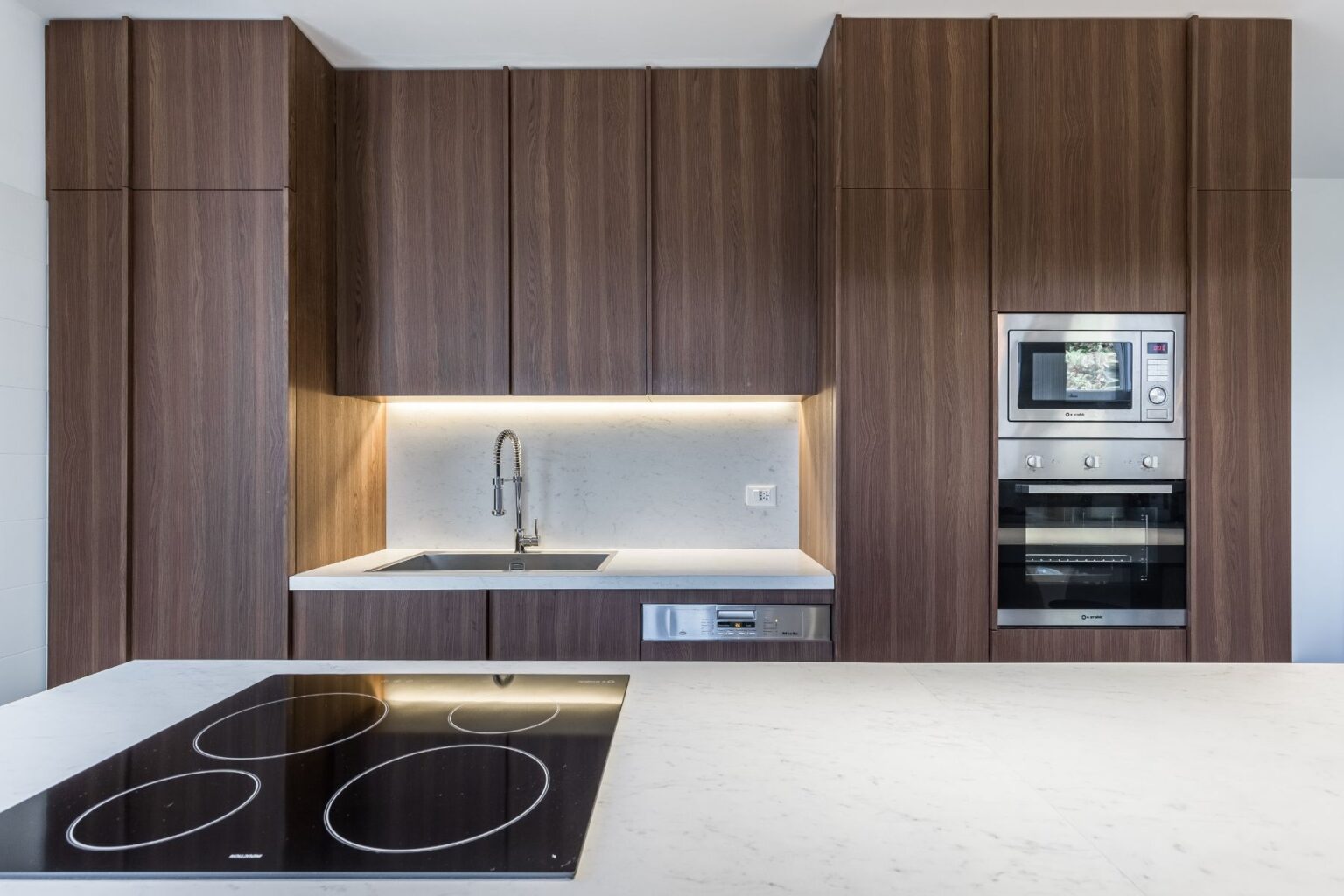 Modern kitchen with dark wood cabinetry, a sleek marble countertop, stainless steel appliances, and an integrated sink and stovetop. The design features under-cabinet lighting, enhancing the contemporary aesthetic. | Only on SopranoVillas