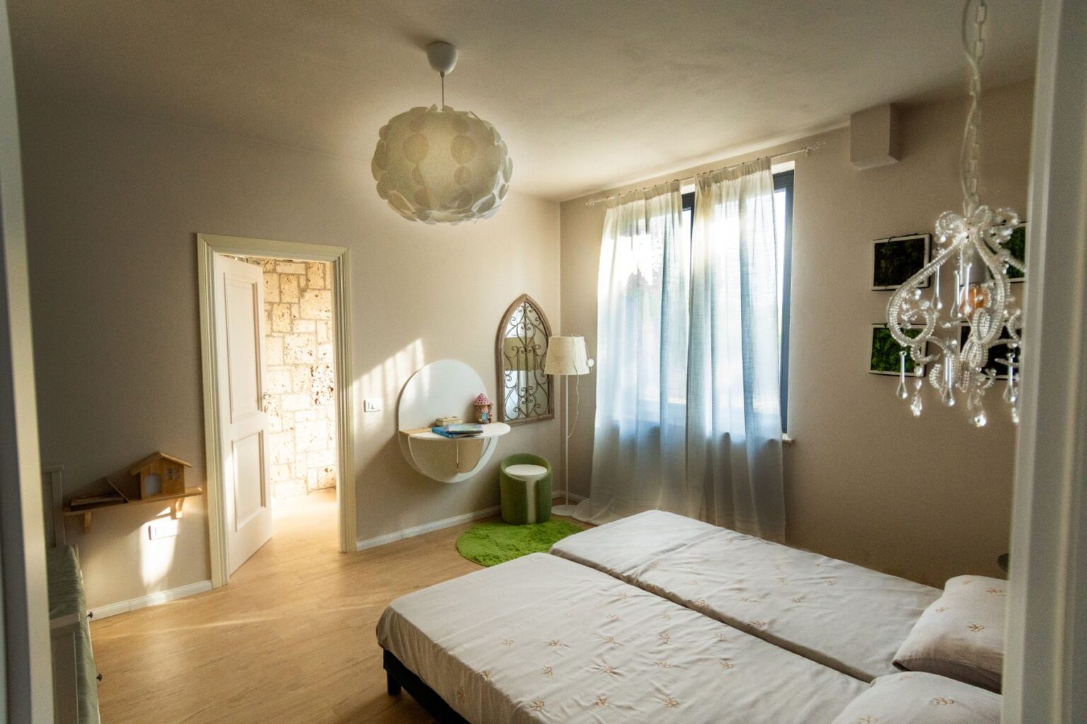 Bright and airy bedroom featuring twin beds with light bedding, a stylish chandelier, a round wall mirror, and a cozy green rug. The room is decorated with sheer curtains allowing natural light to fill the space, creating a warm and inviting atmosphere. | Only on SopranoVillas