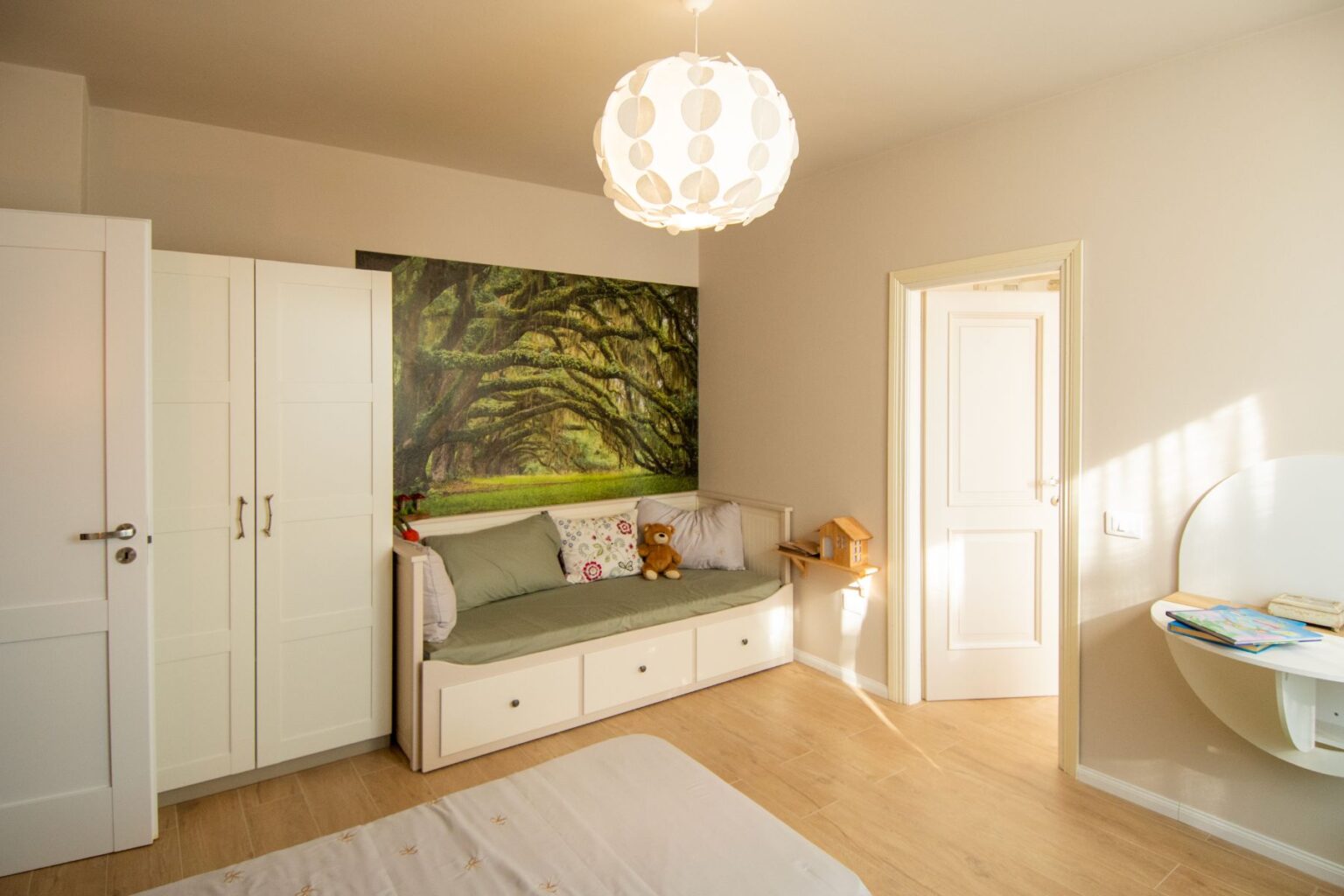 Cozy children's bedroom featuring a green-themed accent wall with a nature mural, a comfortable daybed with pillows, a white wardrobe, and a small wooden shelf with a toy house. The room is illuminated by a modern pendant light, creating a warm and inviting atmosphere. | Only on SopranoVillas