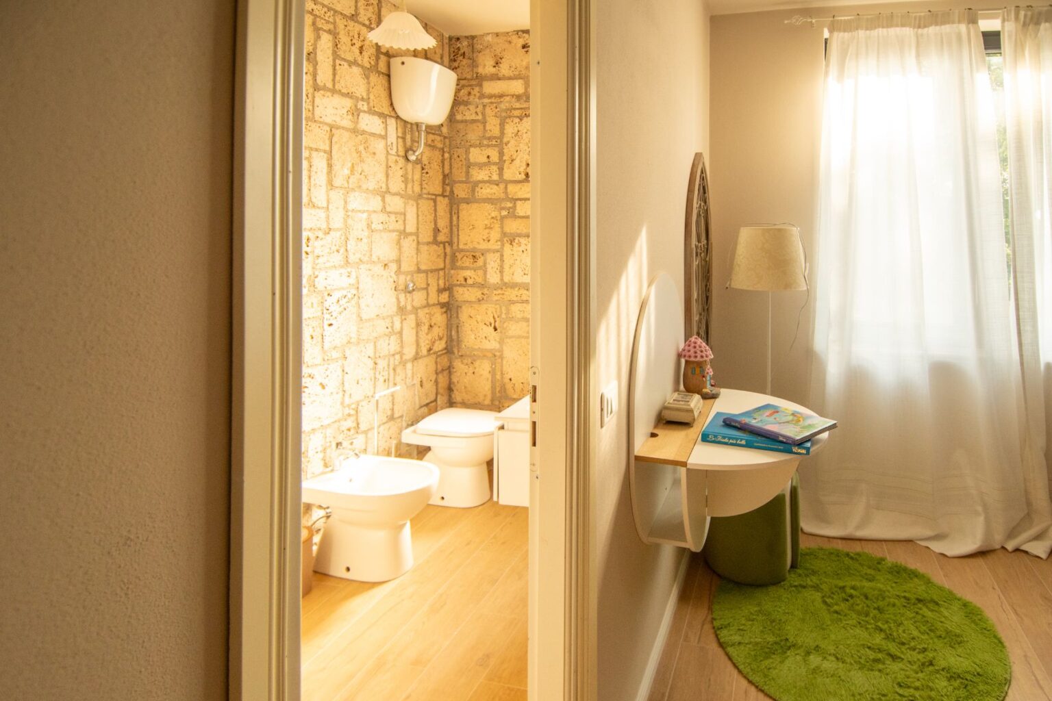 Interior view of a modern bathroom and adjacent room featuring a stone accent wall, a bidet, and a toilet. The room includes a cozy workspace with a round table, books, a decorative lamp, and a green rug, illuminated by natural light from a nearby window. | Only on SopranoVillas