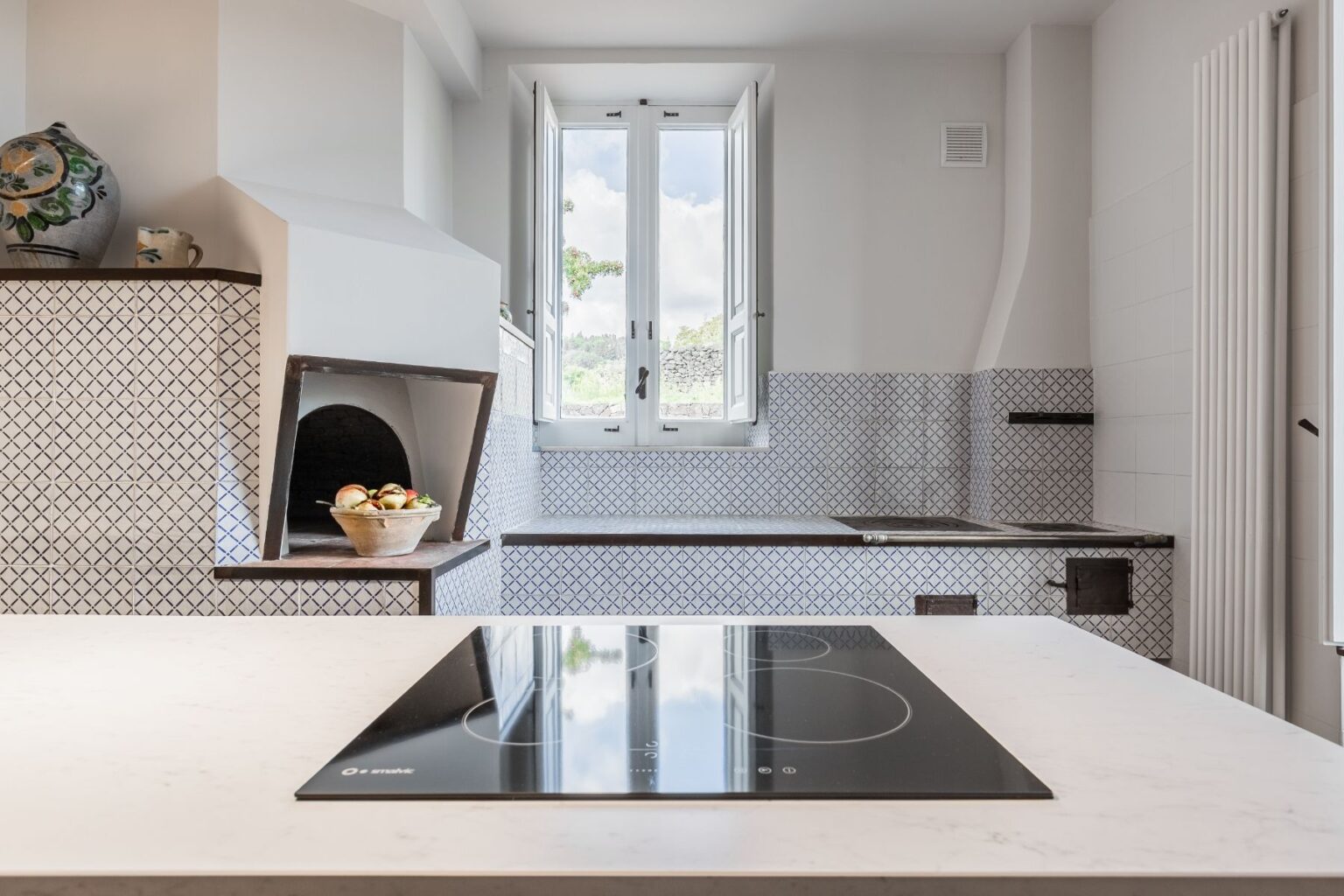 Modern kitchen interior featuring a sleek induction cooktop, white marble countertop, and a charming traditional pizza oven. Natural light floods in through double windows, illuminating the stylish blue and white tiled backsplash and minimalistic design elements. Ideal for contemporary cooking and entertaining. | Only on SopranoVillas