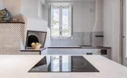 Modern kitchen interior featuring a sleek induction cooktop, white marble countertop, and a charming traditional pizza oven. Natural light floods in through double windows, illuminating the stylish blue and white tiled backsplash and minimalistic design elements. Ideal for contemporary cooking and entertaining.