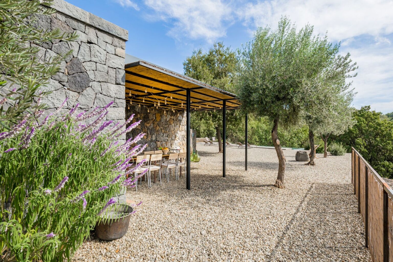 Modern outdoor dining area with a stone wall, surrounded by lush greenery and flowering plants. The space features a covered pergola with wooden beams and metal supports, offering a relaxing atmosphere for meals in nature. | Only on SopranoVillas