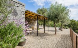 Modern outdoor dining area with a stone wall, surrounded by lush greenery and flowering plants. The space features a covered pergola with wooden beams and metal supports, offering a relaxing atmosphere for meals in nature.