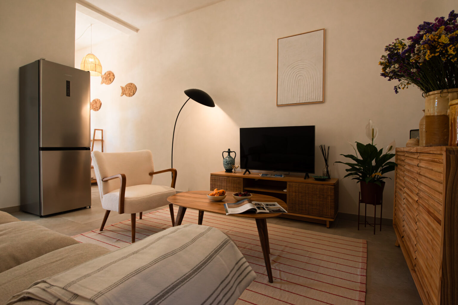 Cozy living room featuring a modern design with a beige armchair, wooden coffee table, and a sleek television on a media console. A stainless steel refrigerator is visible in the background, complemented by decorative fish wall art and a stylish floor lamp. Fresh flowers in a vase and a plant add a touch of nature to the space. The room is illuminated by natural light, creating a warm and inviting atmosphere. | Only on SopranoVillas