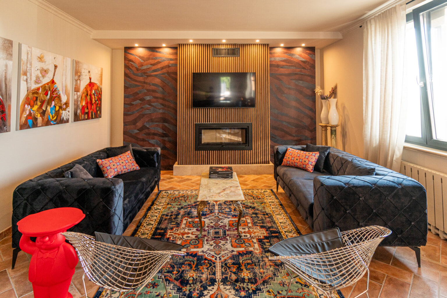 Modern living room featuring plush black sofas with decorative pillows, a stylish coffee table, and a vibrant area rug. The wall includes a mounted flat-screen TV and a contemporary fireplace, accented by artistic wall decor. A unique red sculpture and wire chairs add a playful touch to the elegant space. Natural light streams in through large windows, enhancing the inviting atmosphere. | Only on SopranoVillas